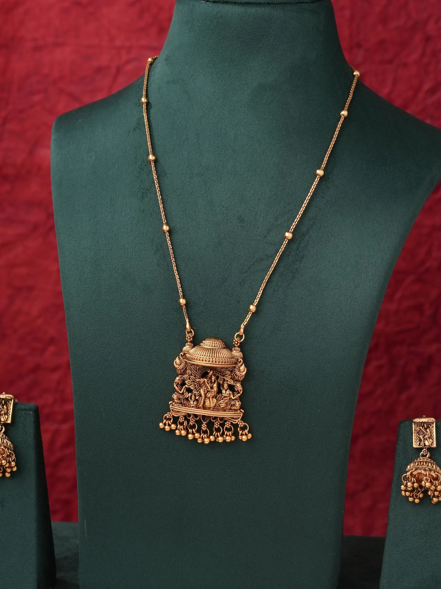 An ornate gold-toned necklace with a detailed pendant displayed on a dark green jewelry bust, with matching earrings partially visible on side stands against a textured red background.