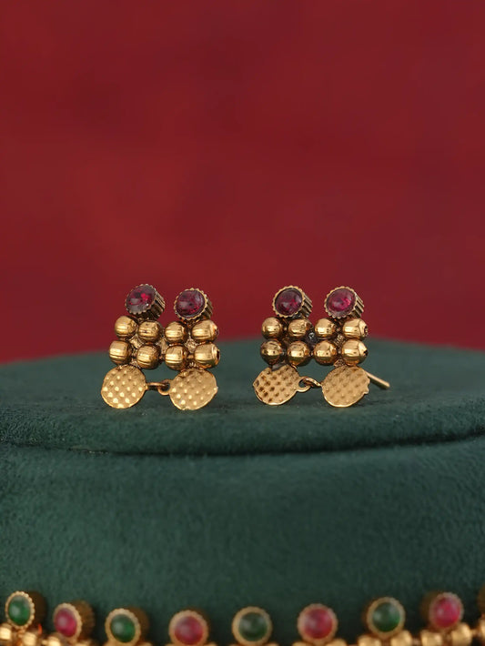 A pair of gold-toned traditional Indian earrings with small gold beads, textured gold disc charms, and round red gemstones at the top, displayed on a dark green velvet surface against a solid red background.