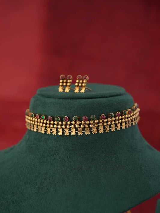 Gold-toned traditional Indian jewelry set featuring a choker necklace and matching earrings, decorated with small red and green stones and gold-toned floral motifs, displayed on a dark green velvet jewelry bust against a dark red background.