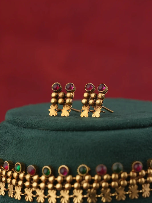 A pair of gold-toned traditional Indian earrings with small bead clusters and circular red gemstones, displayed on a dark green velvet jewelry stand against a deep red background.