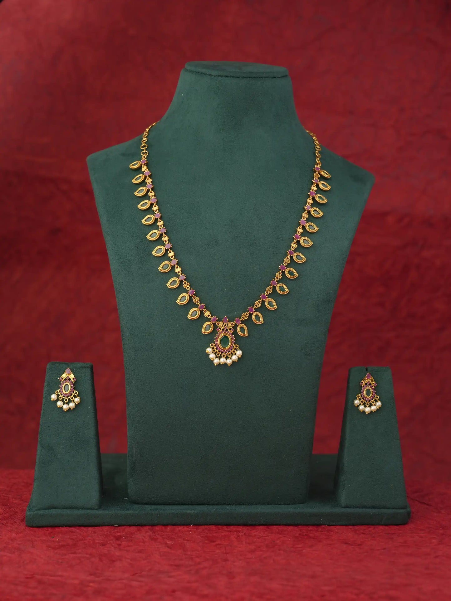 Gold-toned traditional Indian jewelry set featuring a necklace and matching earrings displayed on a dark green velvet jewelry bust against a textured red background.