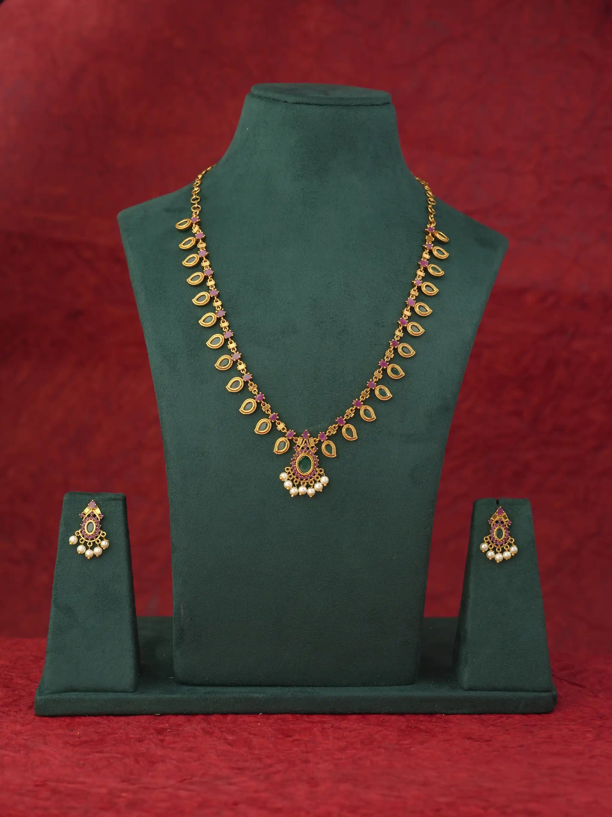 Gold-toned traditional Indian jewelry set featuring a necklace and matching earrings displayed on a dark green velvet jewelry bust against a textured red background.