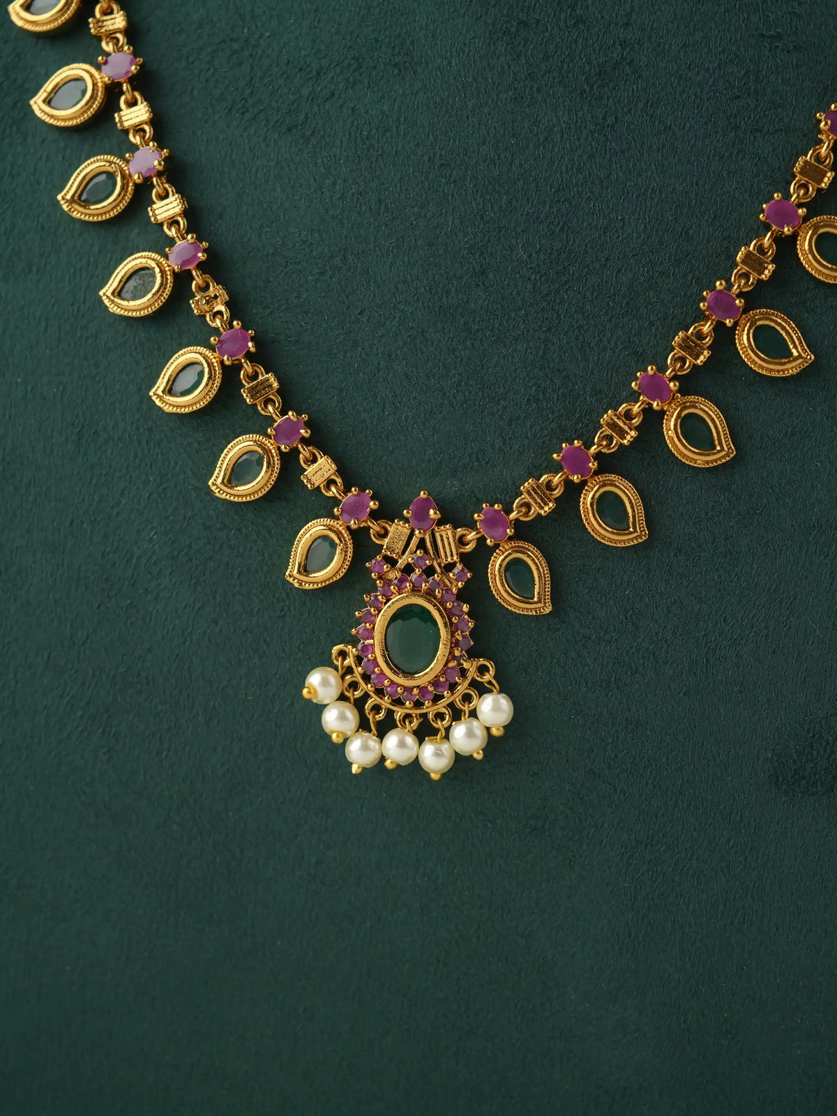 Gold-toned traditional necklace with teardrop-shaped links, purple gemstone accents, and a central pendant decorated with white pearls, displayed on a dark green textured background.