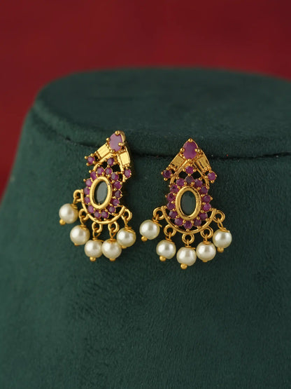 A pair of gold-toned traditional Indian chandelier earrings with purple gemstone accents and white pearl drops, displayed on a dark green velvet jewelry stand against a red background.