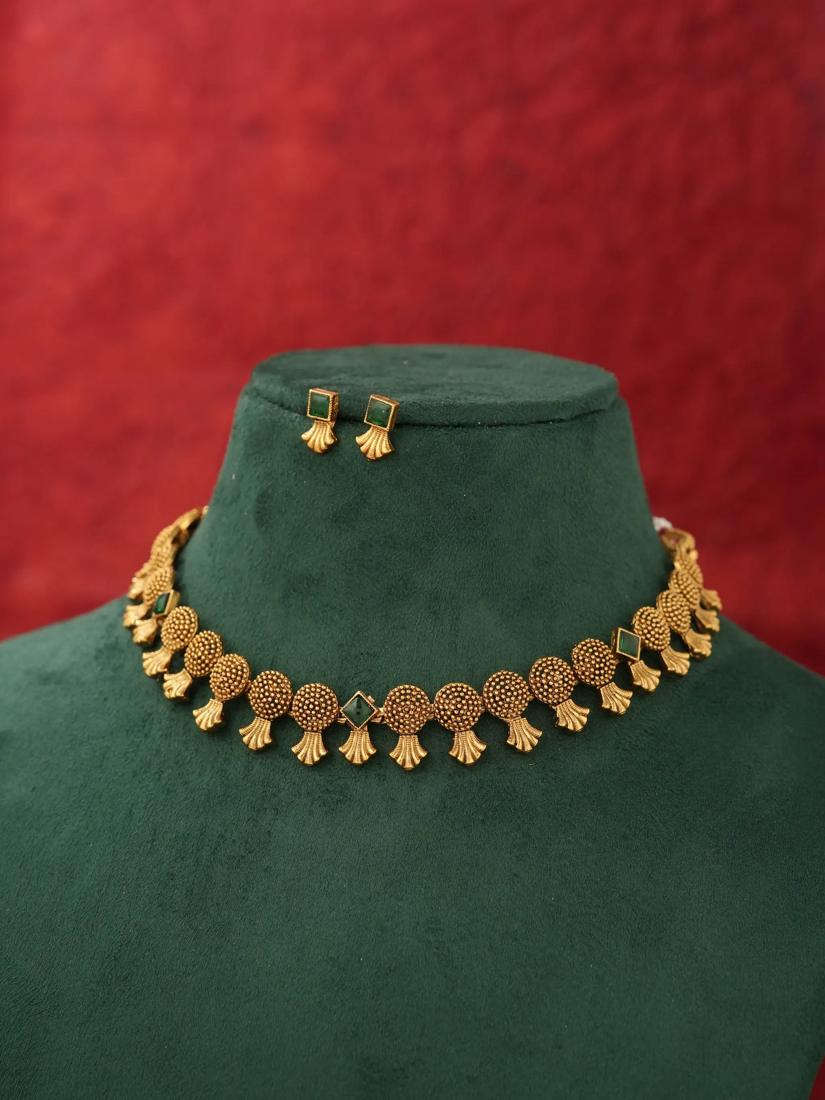Gold-toned traditional necklace and matching rectangular stud earrings displayed on a dark green velvet jewelry bust against a textured red background.