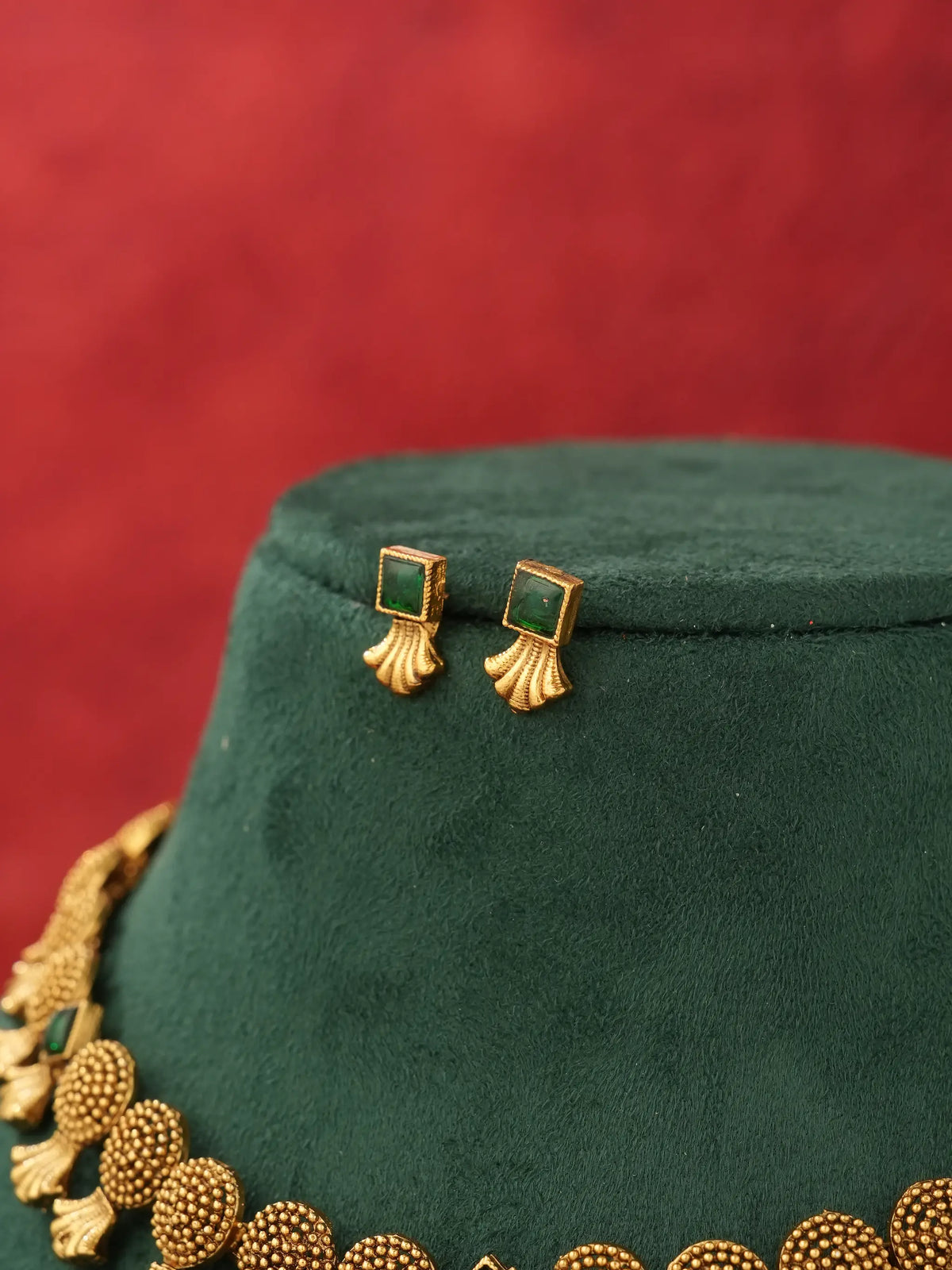 Gold-tone statement earrings with square green gemstones and a matching beaded necklace displayed on a dark green velvet jewelry bust against a textured red background.
