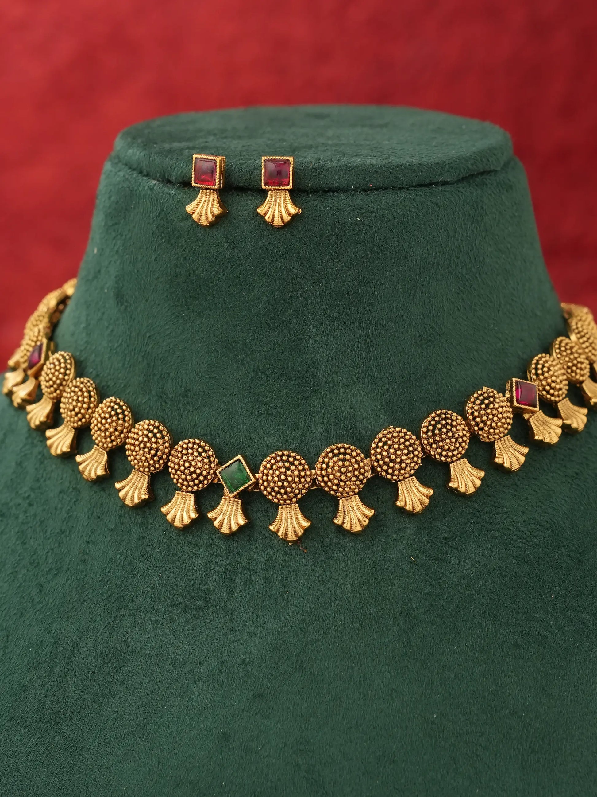 Gold-toned traditional necklace and matching stud earrings displayed on a dark green velvet jewelry bust against a red background.