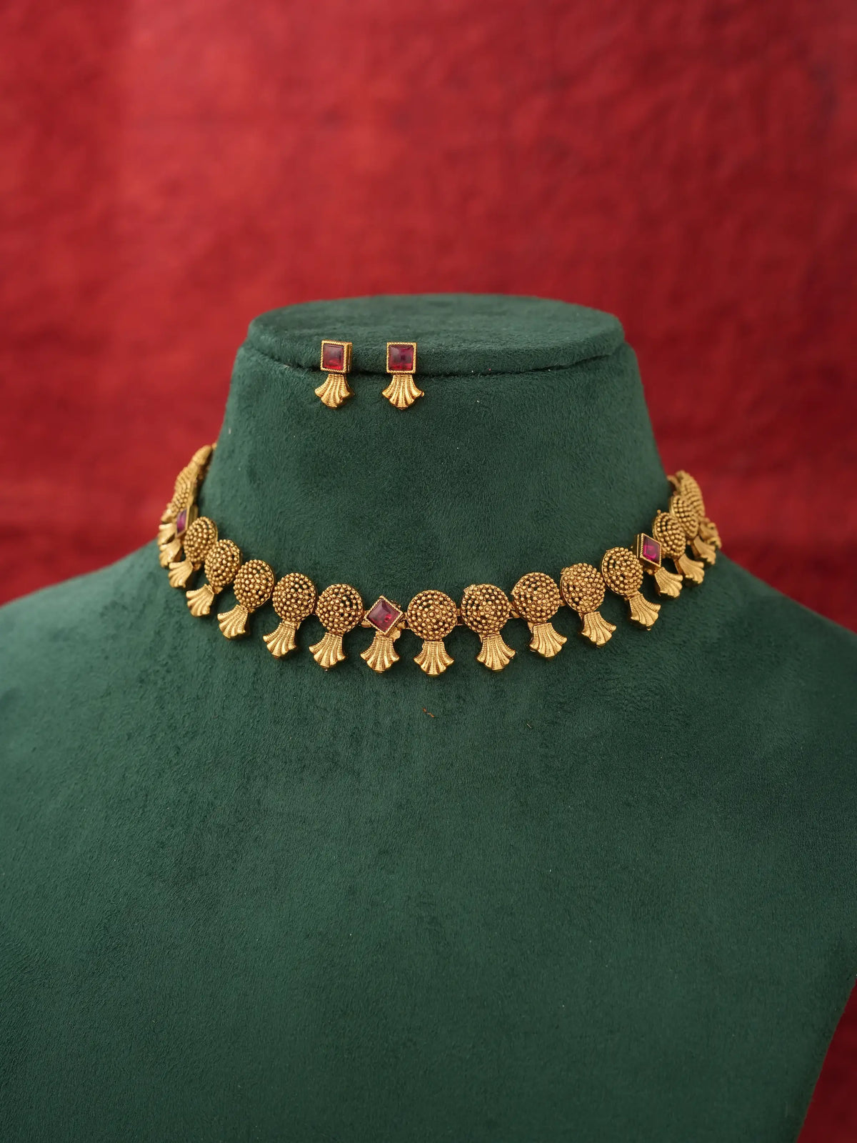 Gold-toned traditional necklace and matching stud earrings displayed on a dark green velvet jewelry bust against a textured red background.