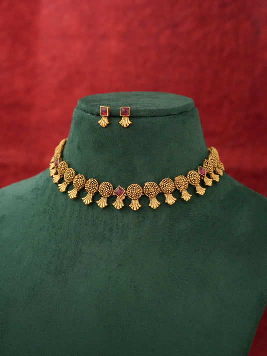 Gold-toned traditional necklace and matching stud earrings displayed on a dark green velvet jewelry bust against a textured red background.