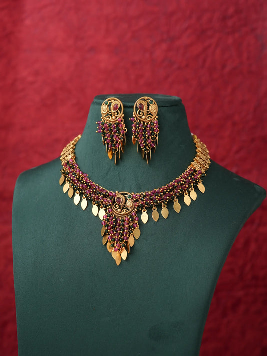 Gold-toned traditional jewelry set featuring a statement necklace and matching drop earrings, embellished with red stones and gold leaf-shaped charms, displayed on a dark green jewelry bust against a textured red background.