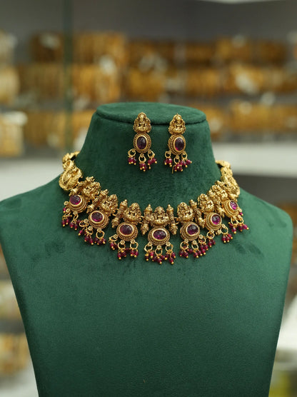 An ornate gold-toned traditional necklace and matching drop earrings displayed on a dark green velvet jewelry bust. The jewelry features circular purple stones and dangling red bead accents, with a softly blurred shop interior in the background.