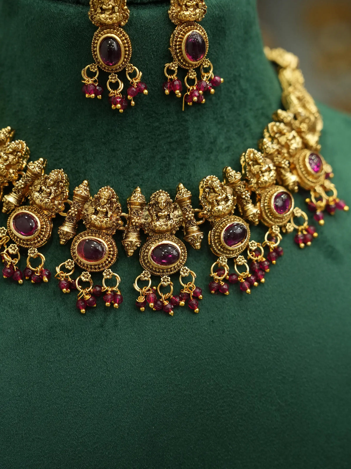 An ornate gold-toned traditional necklace with matching earrings displayed on a dark green velvet jewelry bust. The jewelry features intricate filigree detailing, round purple gemstone centers, and dangling red bead chains.