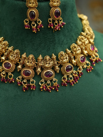 An ornate gold-toned traditional necklace with matching earrings displayed on a dark green velvet jewelry bust. The jewelry features intricate filigree detailing, round purple gemstone centers, and dangling red bead chains.