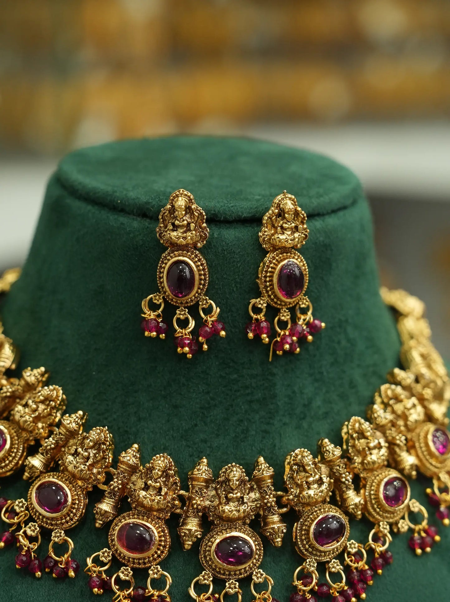 An ornate gold-toned jewelry set displayed on a dark green velvet stand, featuring a matching necklace and earrings with oval purple gemstones and dangling red bead accents.
