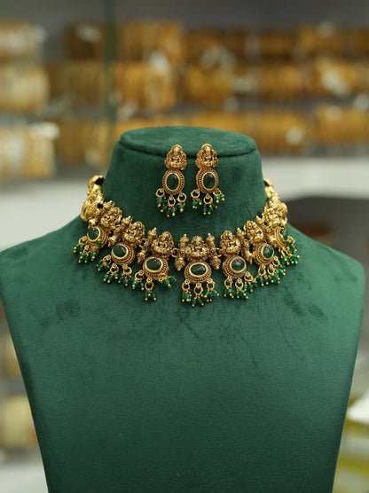 An ornate gold-toned traditional necklace and matching earrings displayed on a dark green velvet jewelry bust. The jewelry features intricate filigree detailing, green stone accents, and dangling green bead chains, set against a softly blurred background of a jewelry store.