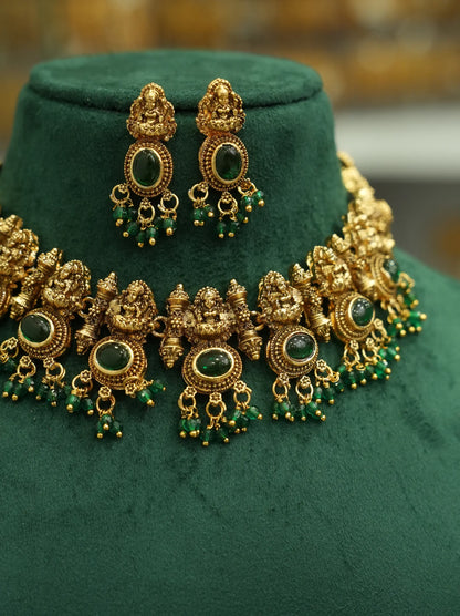 An ornate gold-toned traditional necklace and matching drop earrings displayed on a dark green velvet jewelry bust. The jewelry features detailed filigree, green oval stones, and dangling green bead accents.