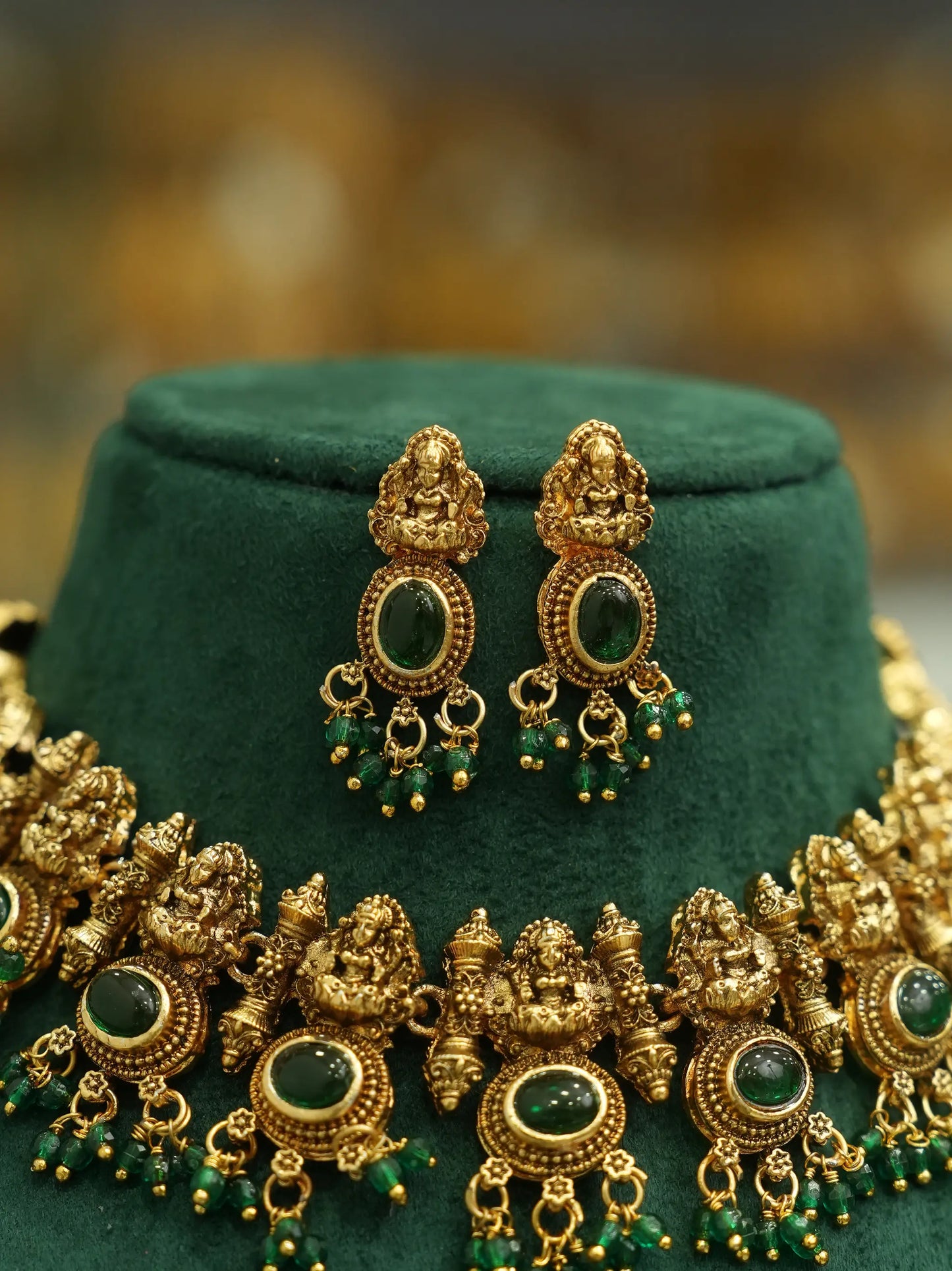 An ornate gold-toned traditional jewelry set displayed on a dark green velvet stand, featuring a necklace and matching earrings with green stone and bead details.