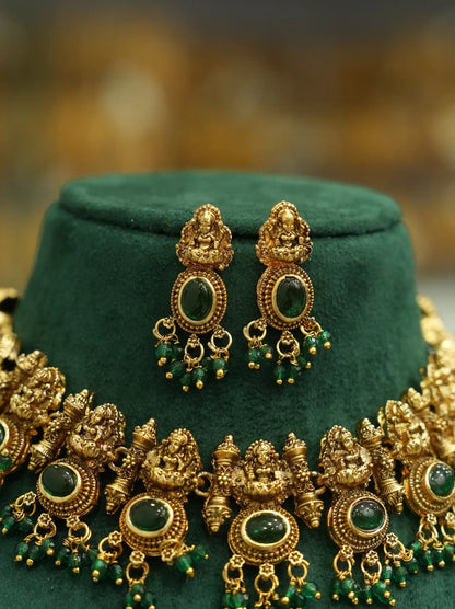 An ornate gold-toned traditional jewelry set displayed on a dark green velvet stand, featuring a necklace and matching earrings with green stone and bead details.