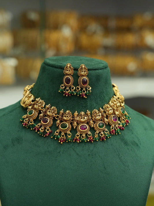 An ornate gold-toned traditional necklace and matching earrings displayed on a dark green velvet jewelry bust. The jewelry features circular purple and green stone accents with dangling red and green bead chains, set against a softly blurred warm-toned background.
