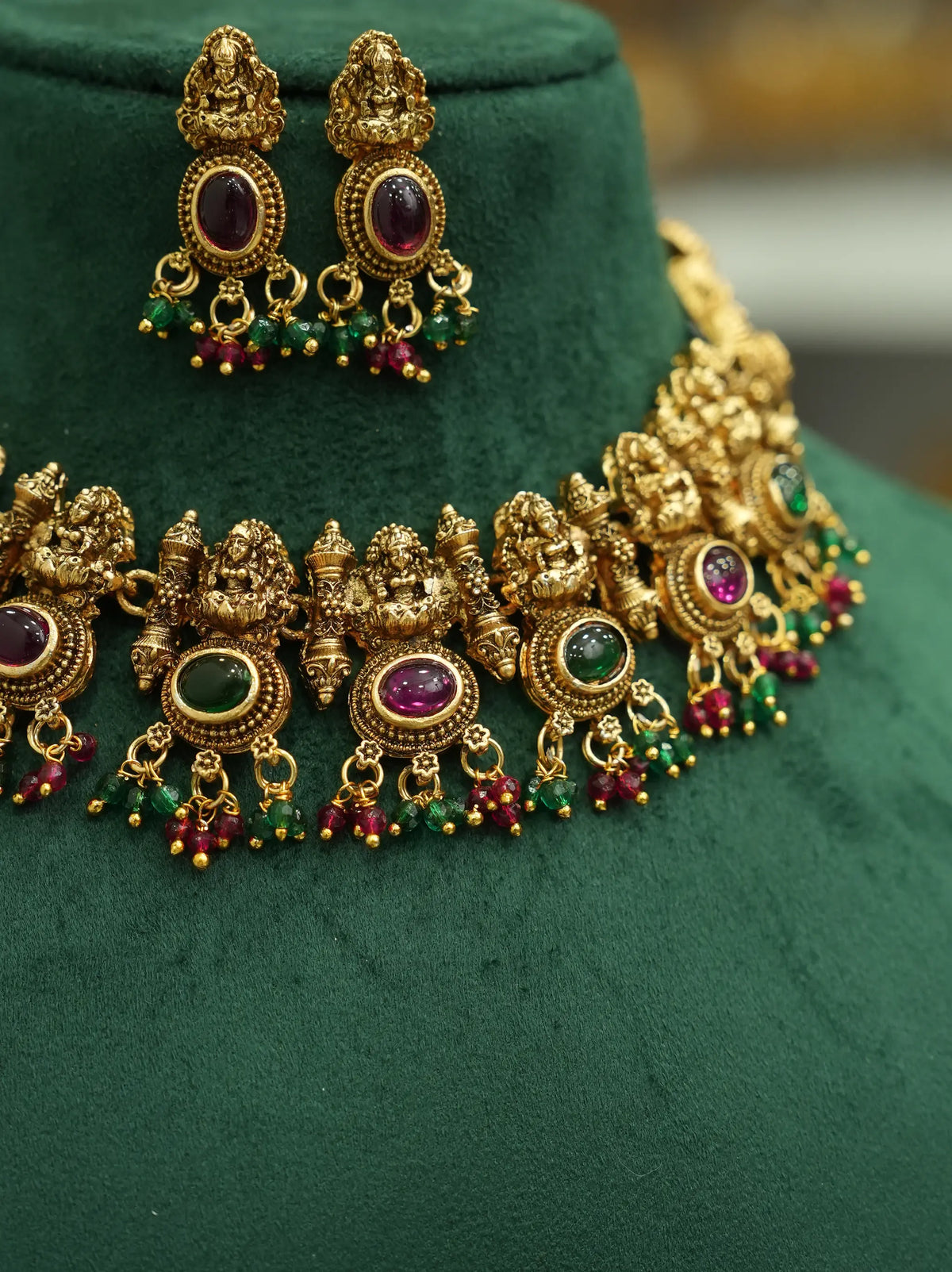 An ornate gold-toned traditional jewelry set displayed on a dark green velvet bust, featuring a matching necklace and earrings with oval purple and green gemstone centers and dangling red and green bead accents.