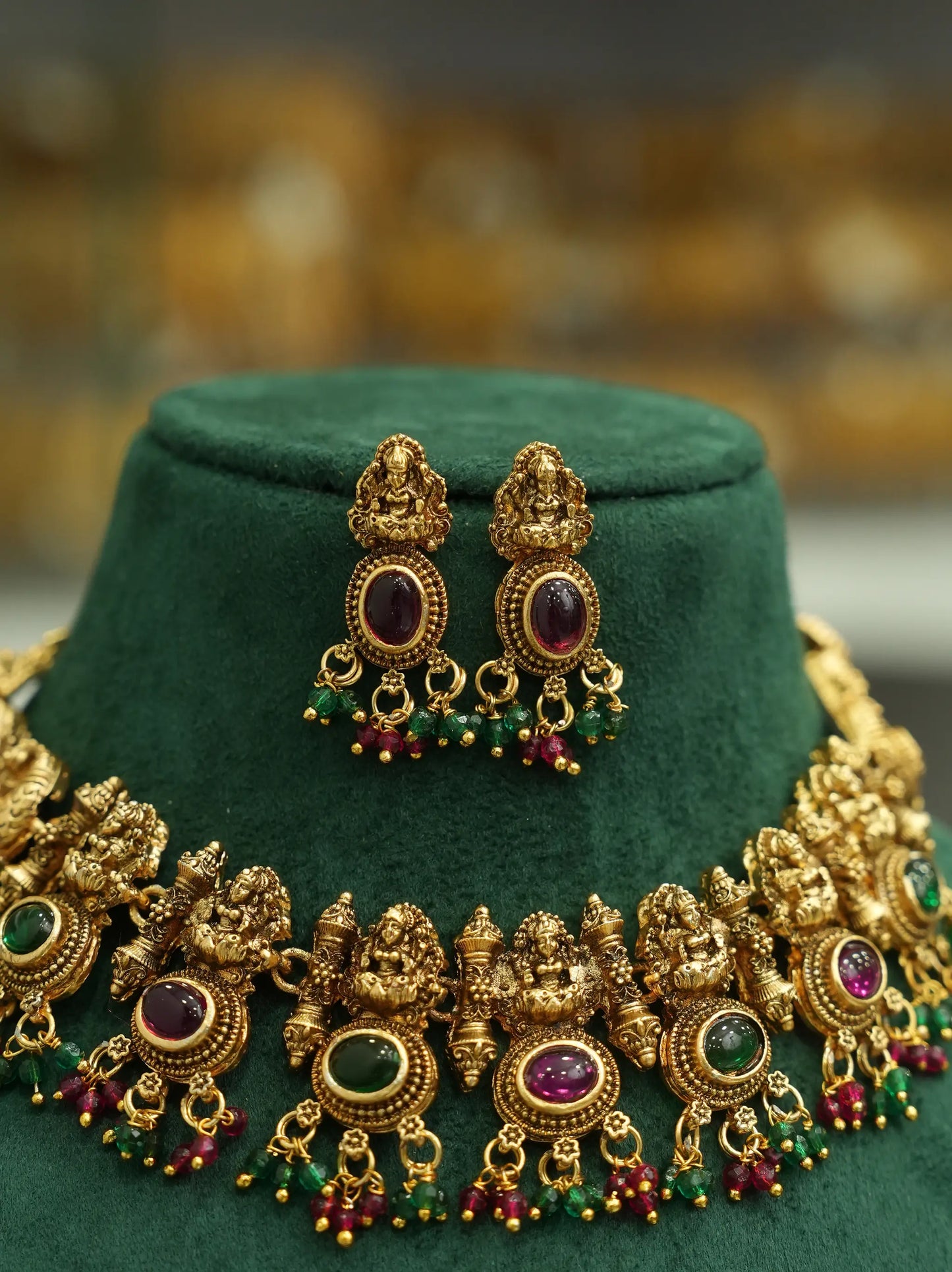 An ornate gold-toned jewelry set displayed on a dark green velvet stand, featuring a matching necklace and earrings with deity motifs and colorful gemstone accents.