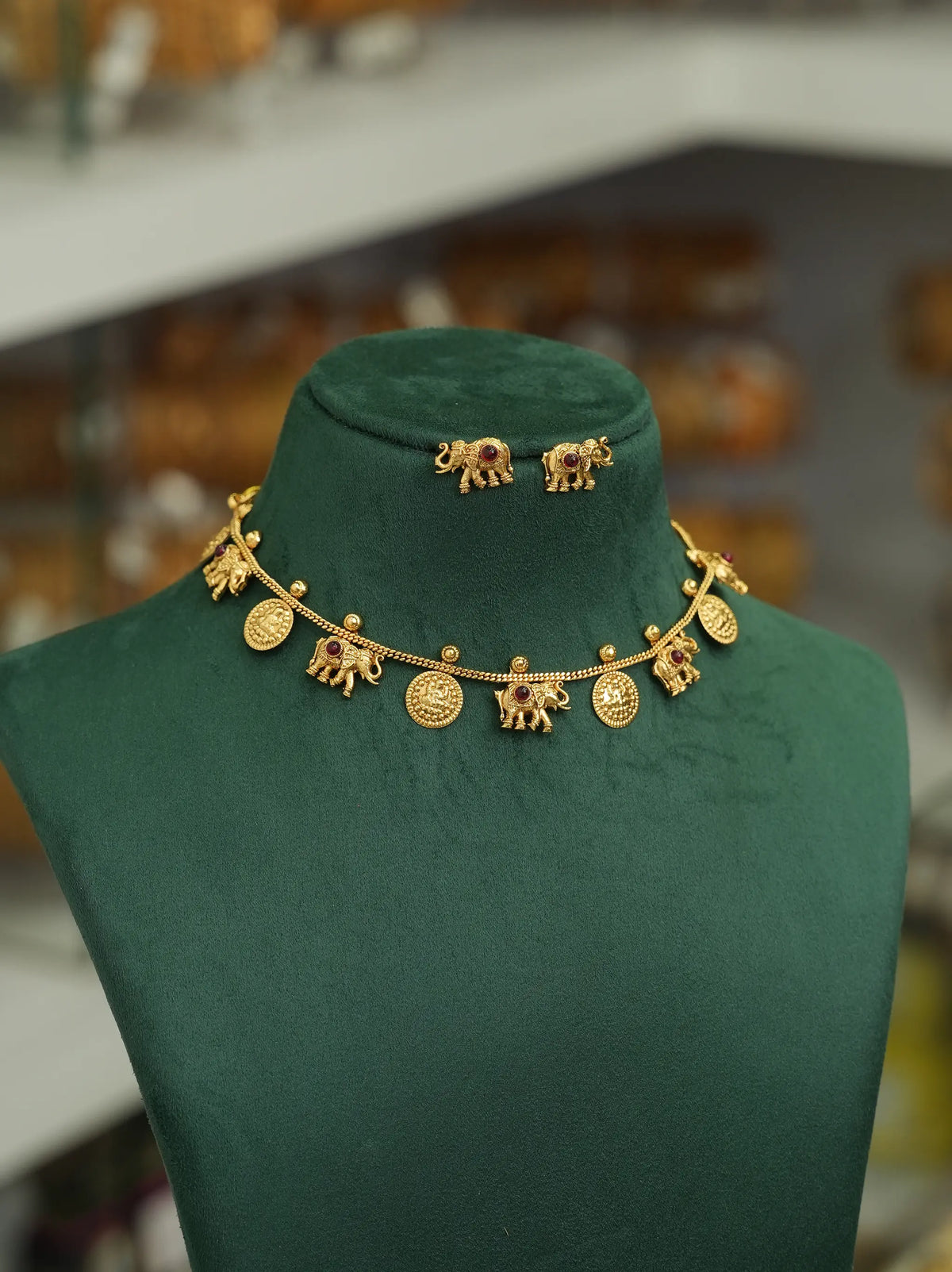 A gold-toned jewelry set displayed on a dark green velvet mannequin bust, featuring a necklace with elephant and coin charms and matching elephant stud earrings.
