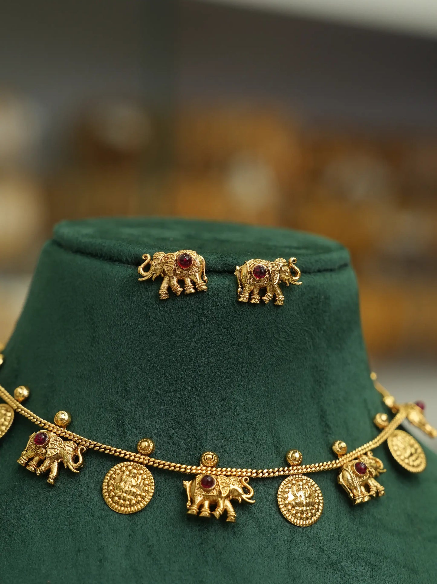 Gold-toned jewelry set displayed on a dark green velvet jewelry bust, featuring elephant-shaped earrings and a matching chain necklace with elephant and coin charms.