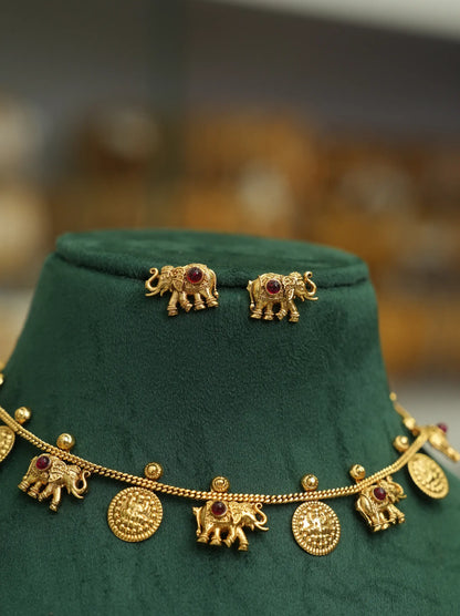 Gold-toned jewelry set displayed on a dark green velvet jewelry bust, featuring elephant-shaped earrings and a matching chain necklace with elephant and coin charms.