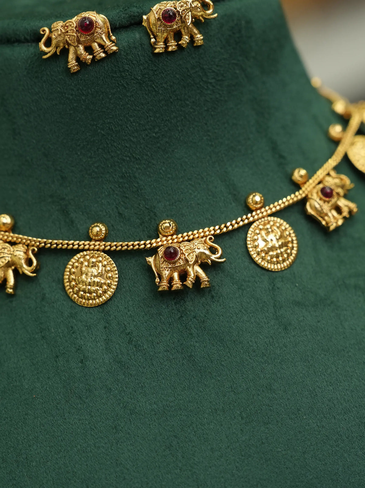 Gold-toned jewelry set featuring elephant-shaped charms and round coin-like discs with small red gemstone accents, displayed on a dark green velvet surface.