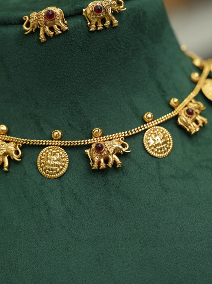 Gold-toned jewelry set featuring elephant-shaped charms and round coin-like discs with small red gemstone accents, displayed on a dark green velvet surface.