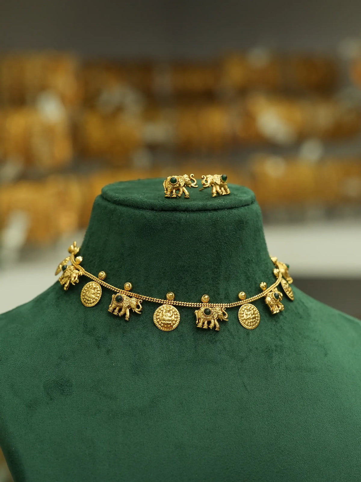 Gold elephant-themed jewelry set displayed on a dark green velvet jewelry bust, featuring a necklace with elephant charms and matching elephant earrings against a blurred background.