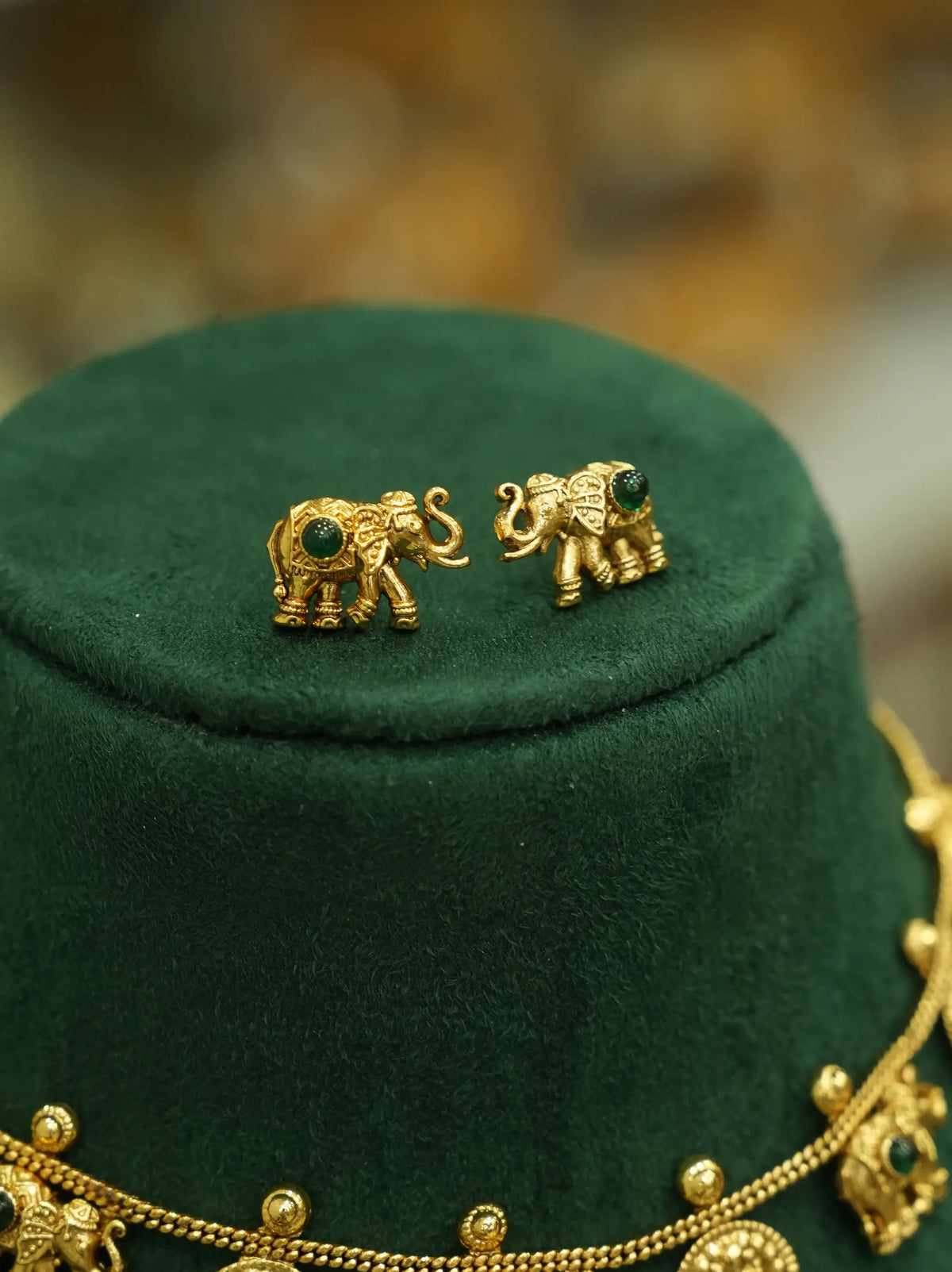 A pair of gold elephant stud earrings with small green stone accents, displayed on a dark green velvet jewelry stand.
