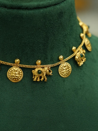 A gold-toned chain necklace displayed on a dark green velvet jewelry bust. The necklace features round textured coin-like charms, a small elephant charm with a dark green stone, and a small bird charm.