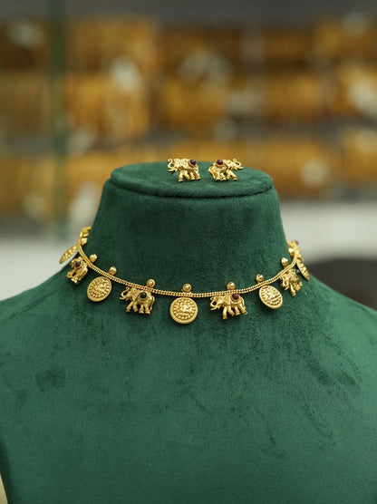 Gold elephant-themed necklace and matching stud earrings displayed on a dark green velvet jewelry bust, with a softly blurred gold-toned shop background.