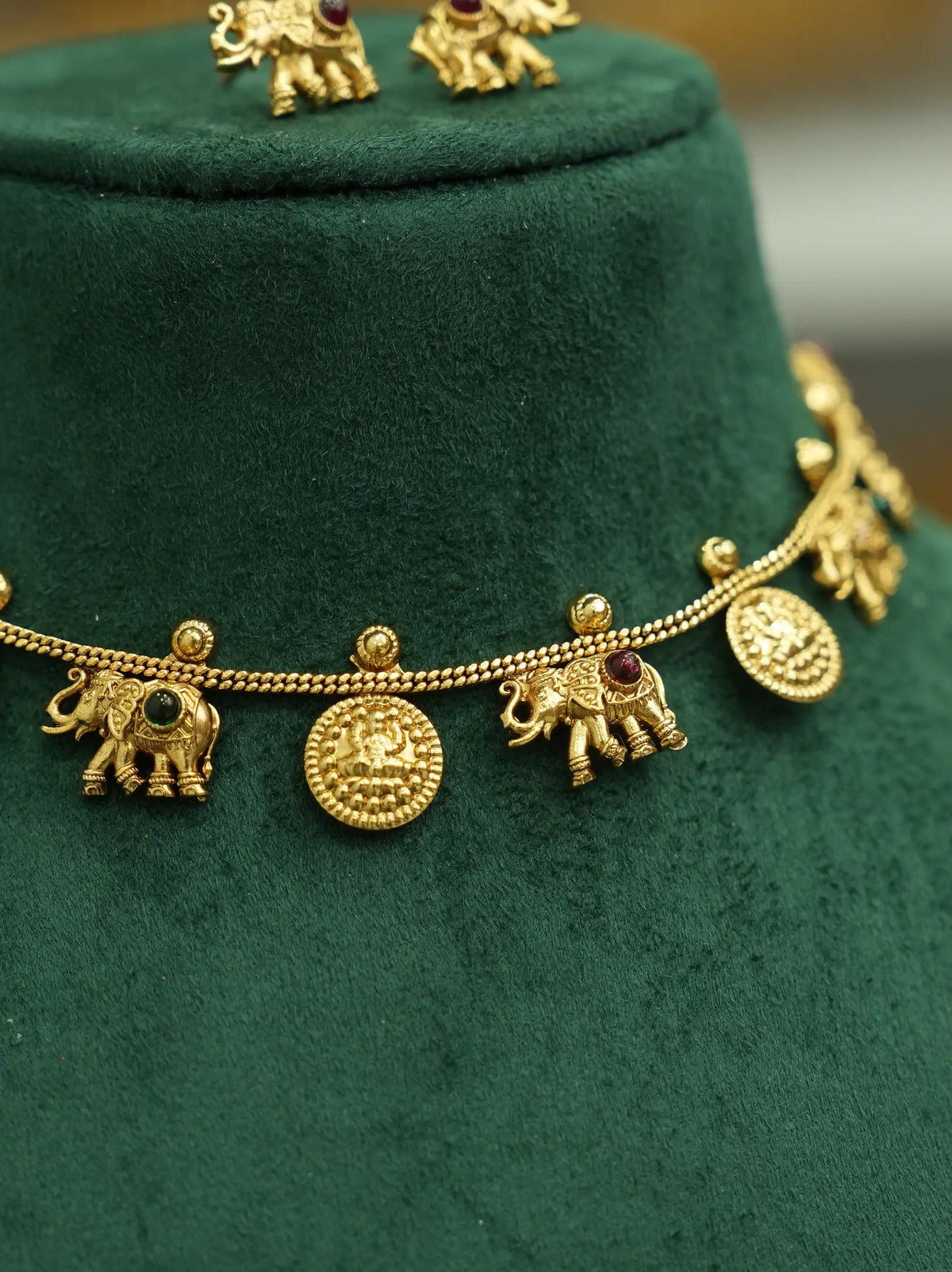 Gold-toned traditional necklace with elephant and coin charms, displayed on a dark green velvet jewelry bust.