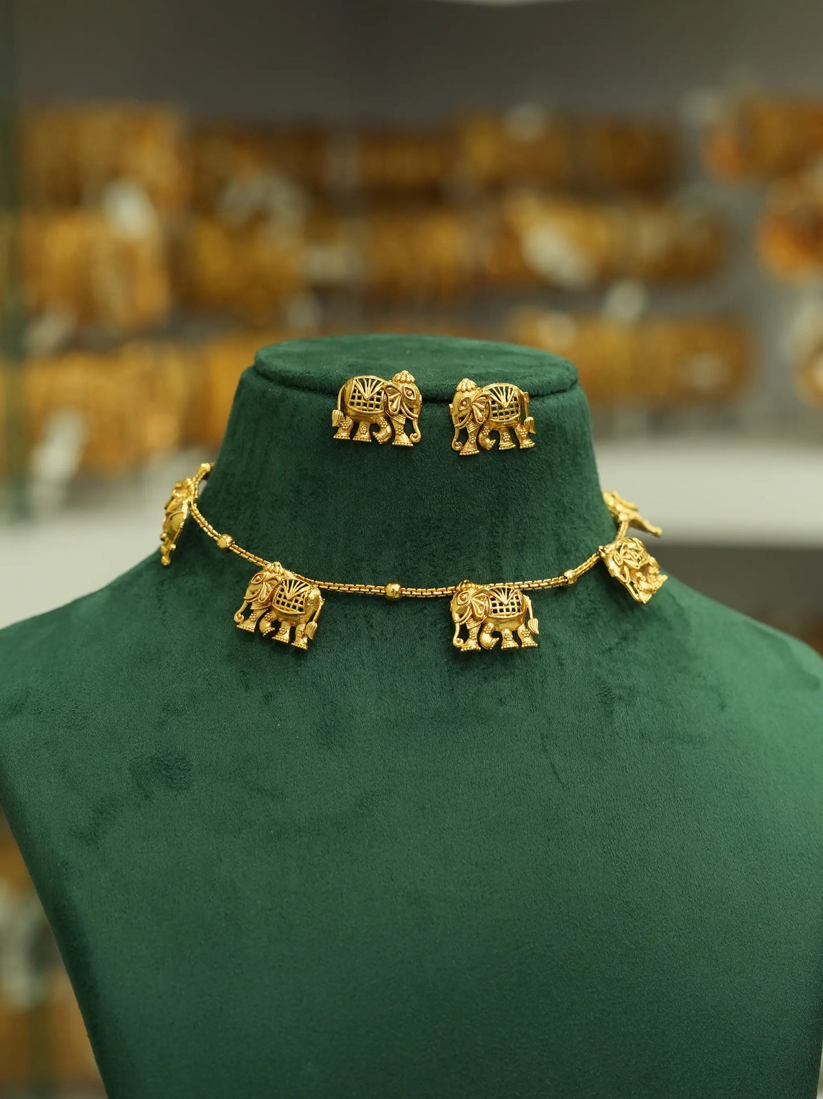 Gold elephant jewelry set including a necklace and matching earrings displayed on a dark green velvet jewelry bust, with a blurred shop background.
