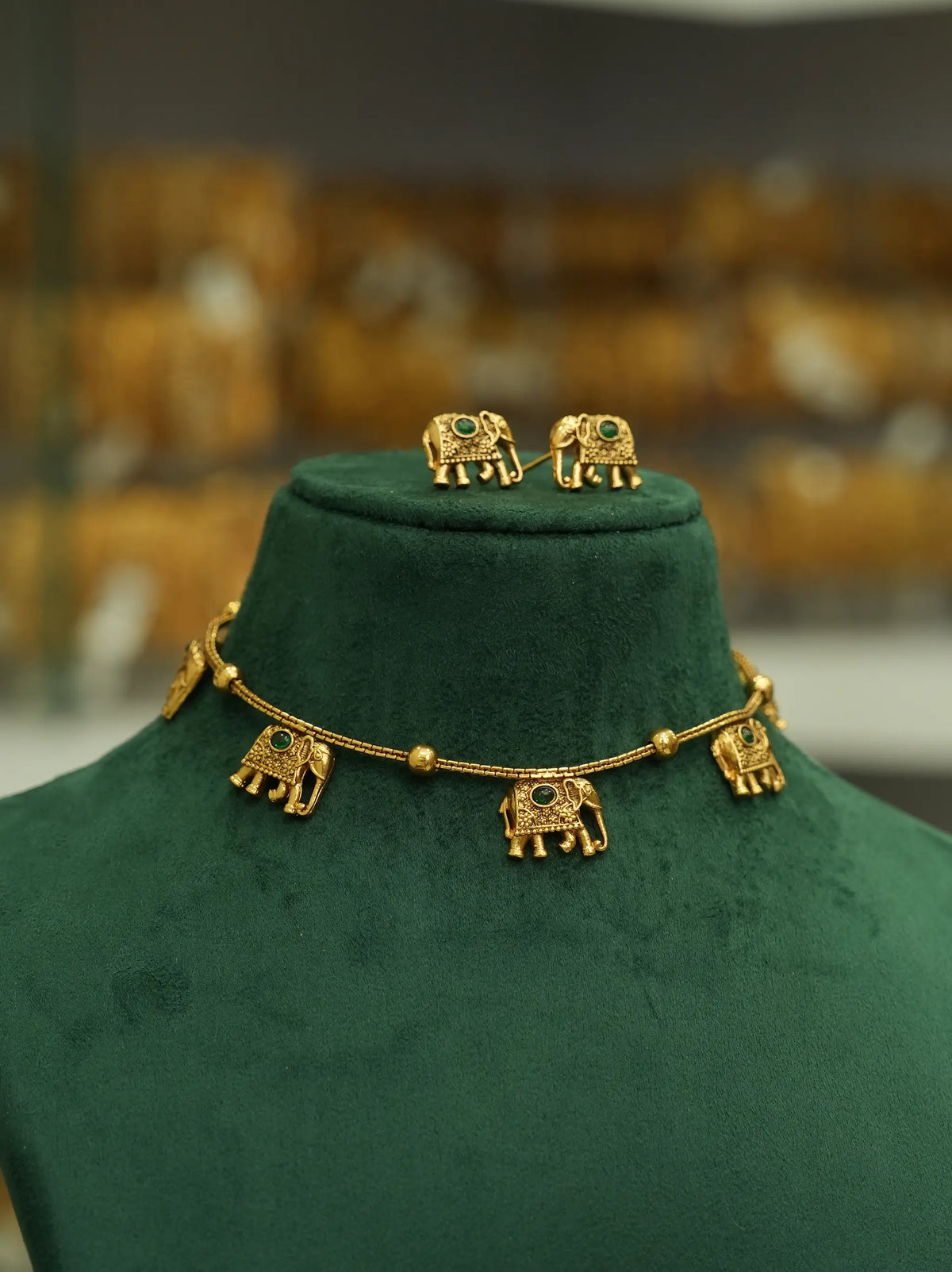 Gold elephant-themed jewelry set displayed on a dark green velvet jewelry bust, featuring a necklace and matching earrings.