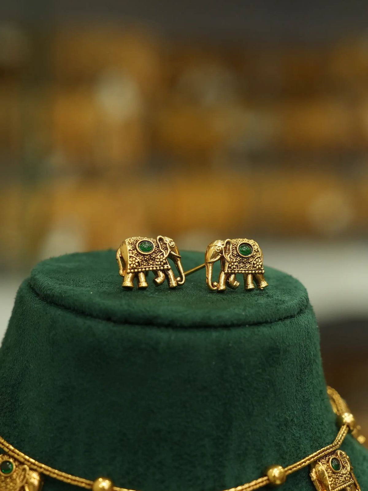 A pair of gold elephant stud earrings with green stone accents, displayed on a dark green velvet jewelry stand.
