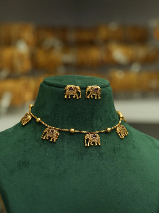 Gold elephant jewelry set featuring a necklace and matching stud earrings displayed on a dark green velvet jewelry bust. The gold pieces are detailed with small red stones and have elephant-shaped charms on the chain.