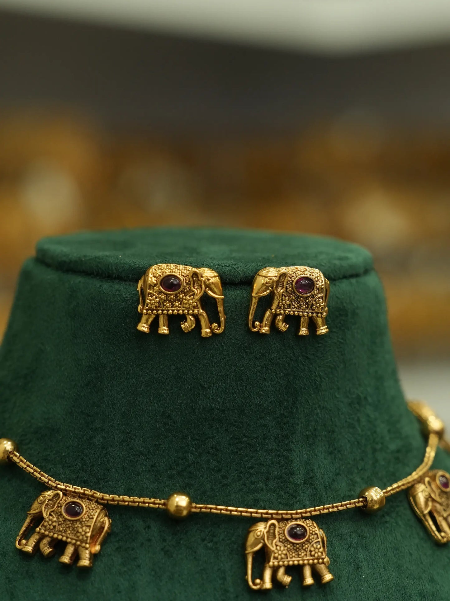 Gold elephant jewelry set displayed on a dark green velvet stand, including a necklace and matching stud earrings with small purple stone accents.