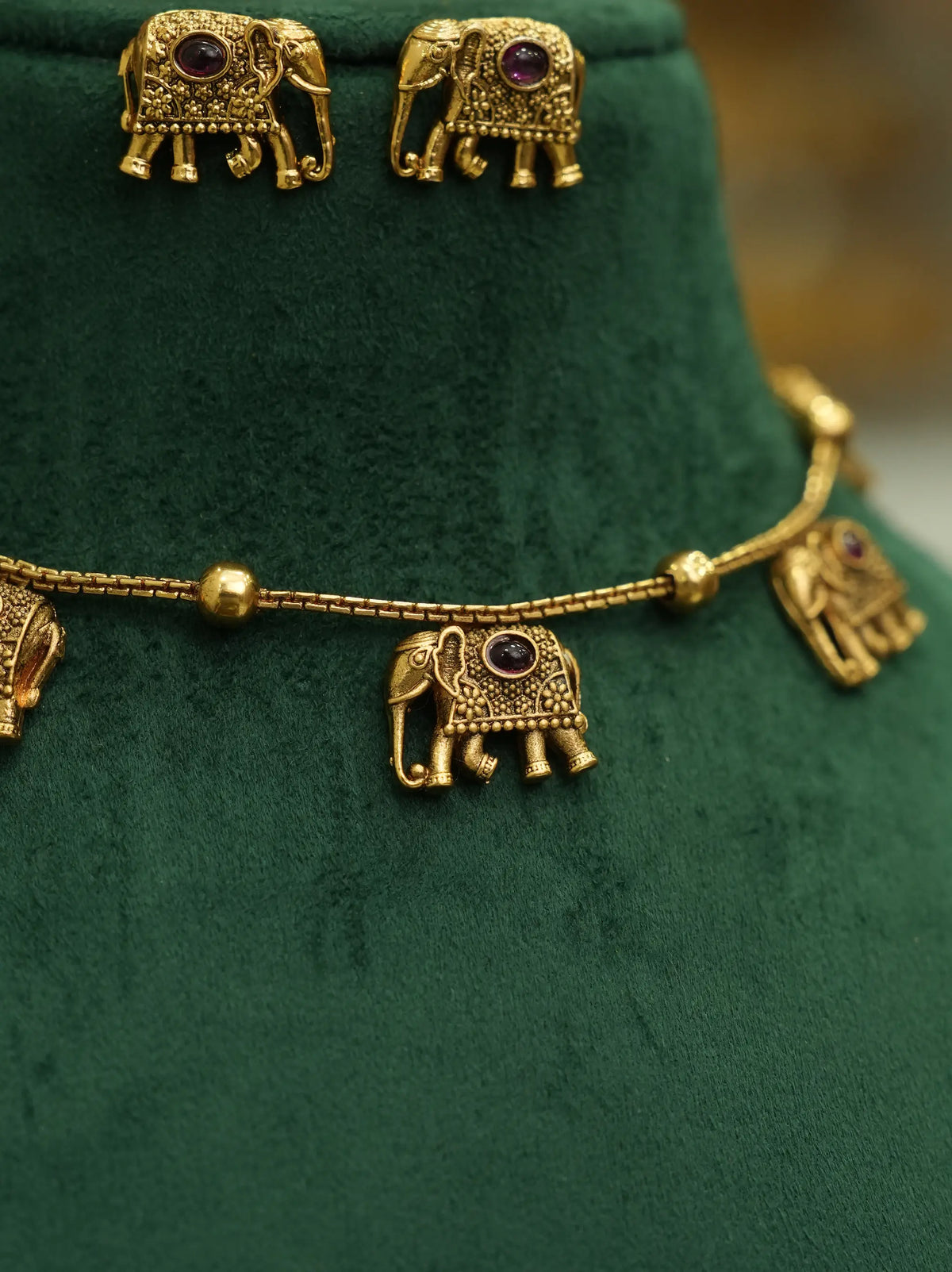 Gold-toned jewelry set featuring elephant-shaped earrings and a matching chain necklace with small elephant charms and round purple stone accents, displayed on a dark green velvet jewelry bust.