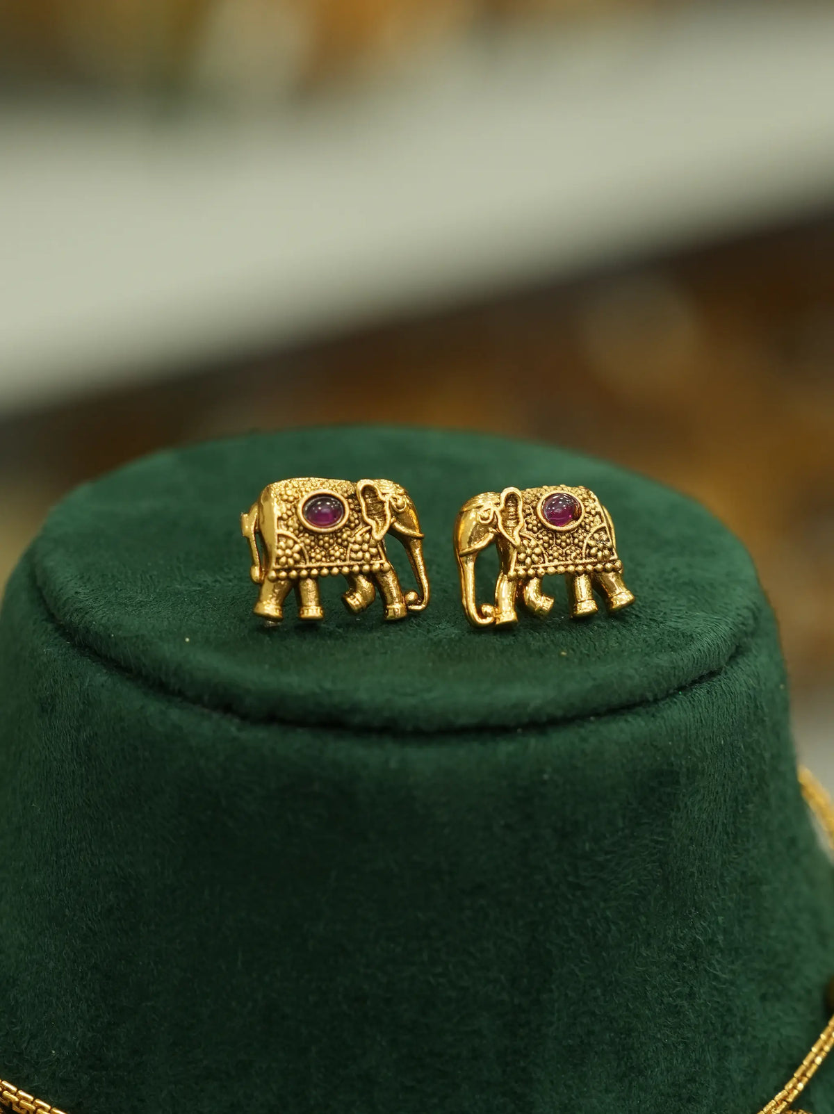 A pair of gold elephant stud earrings with small purple gemstone accents, displayed on a dark green velvet jewelry stand.