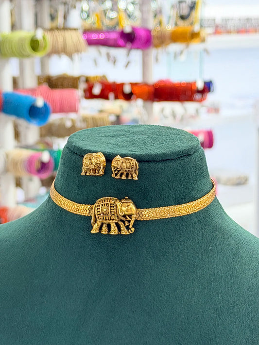 A gold-toned elephant-themed jewelry set displayed on a dark green velvet mannequin bust. The set includes a choker necklace with a central elephant charm, a pair of matching stud earrings, and a small rectangular pendant.