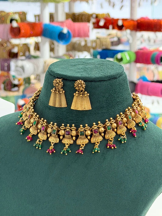 Gold-toned traditional Indian jewelry set featuring a necklace and matching earrings displayed on a dark green velvet mannequin bust, with a colorful blurred jewelry shop background.