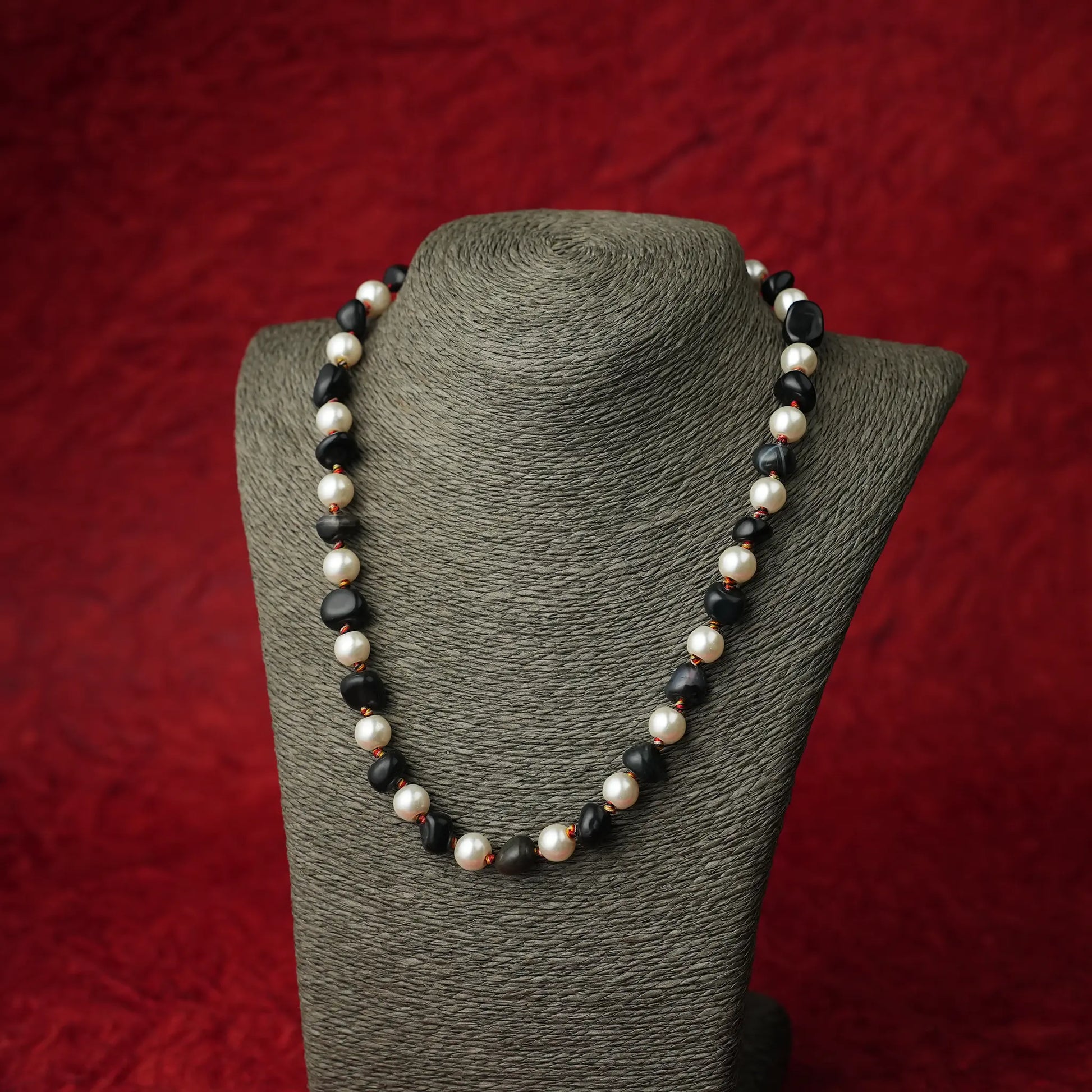 A beaded necklace with alternating white pearls and dark black beads, displayed on a textured gray jewelry bust against a red background.