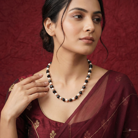 A woman wearing a beaded necklace with black and white pearls, styled with a maroon traditional outfit against a textured dark red background.