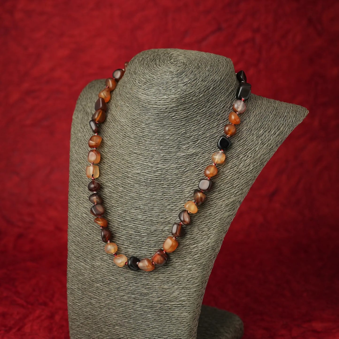 A beaded necklace made of polished irregular gemstone beads in earthy brown, orange, cream, and black tones, displayed on a gray fabric jewelry bust against a textured red background.