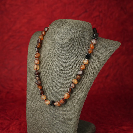 A beaded necklace made of polished irregular gemstone beads in earthy brown, orange, cream, and black tones, displayed on a gray fabric jewelry bust against a textured red background.