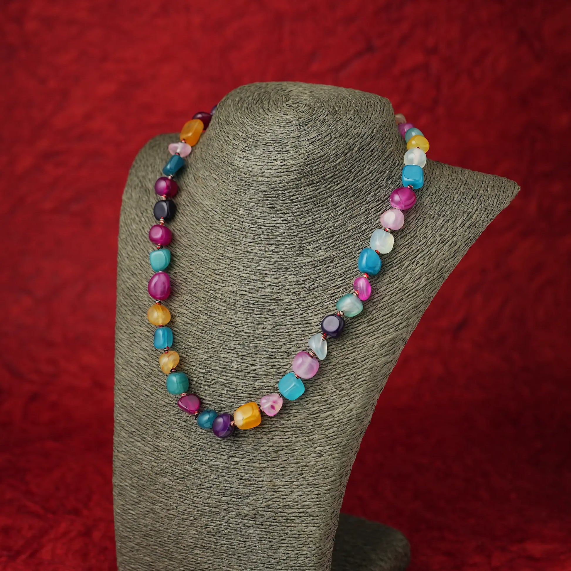 A colorful beaded necklace made of irregular, polished gemstone-like beads in pink, turquoise, purple, yellow, and blue, displayed on a gray fabric jewelry bust against a textured red background.