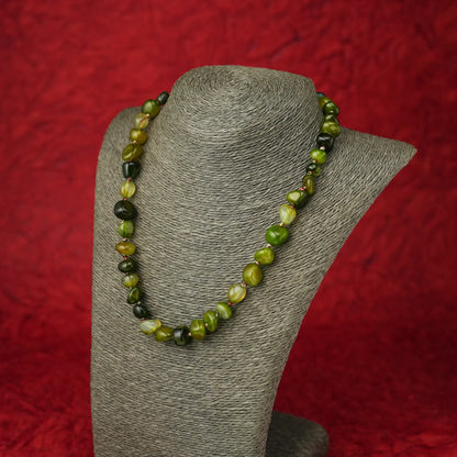 A beaded necklace made of polished olive green and dark forest green marbled beads, displayed on a gray fabric jewelry bust against a textured red background.